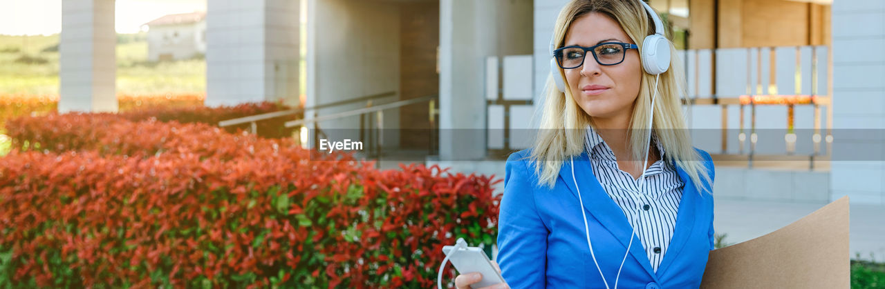 Panoramic view of businesswoman listening music on headphones while standing outdoors