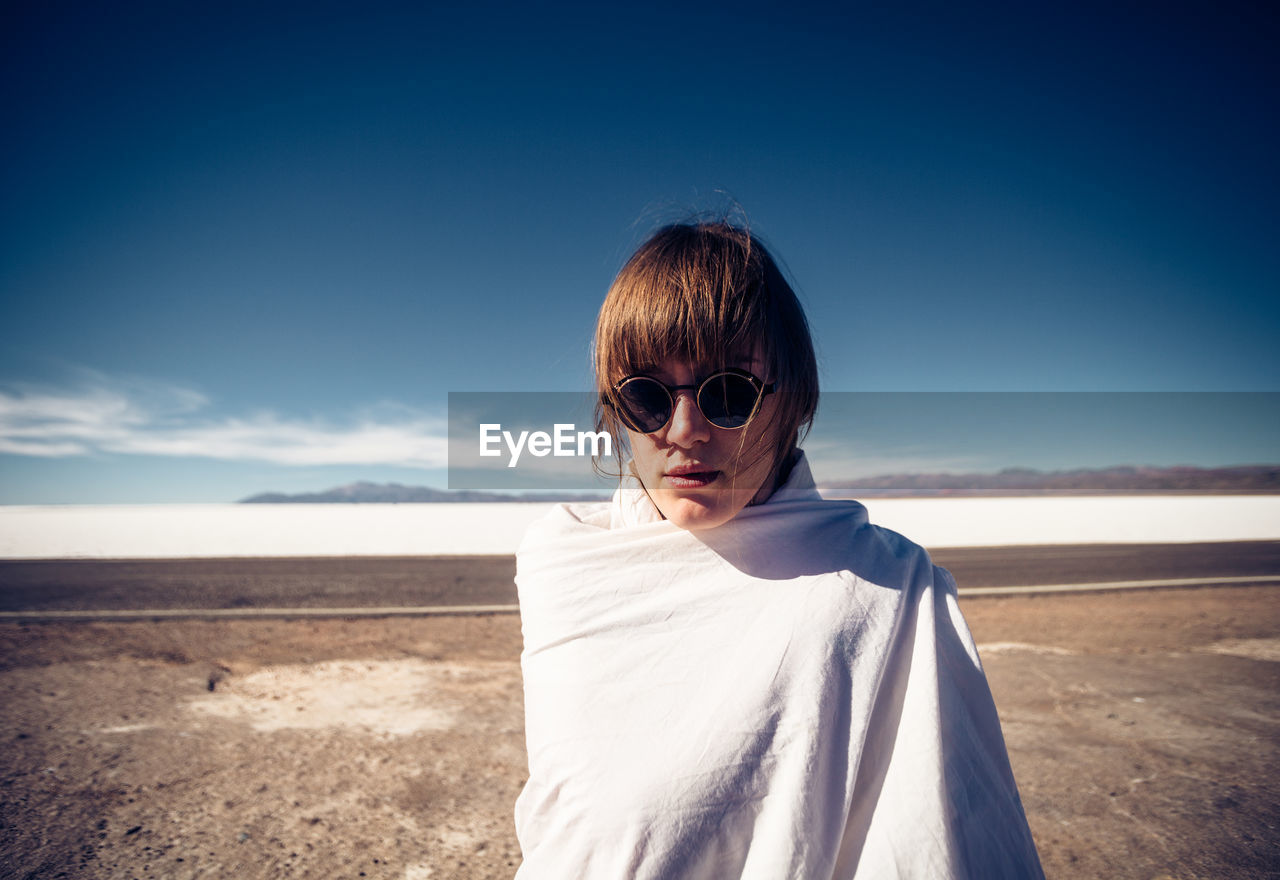 Portrait of woman wearing sunglasses wrapped in blanket standing on land against sky during sunny day