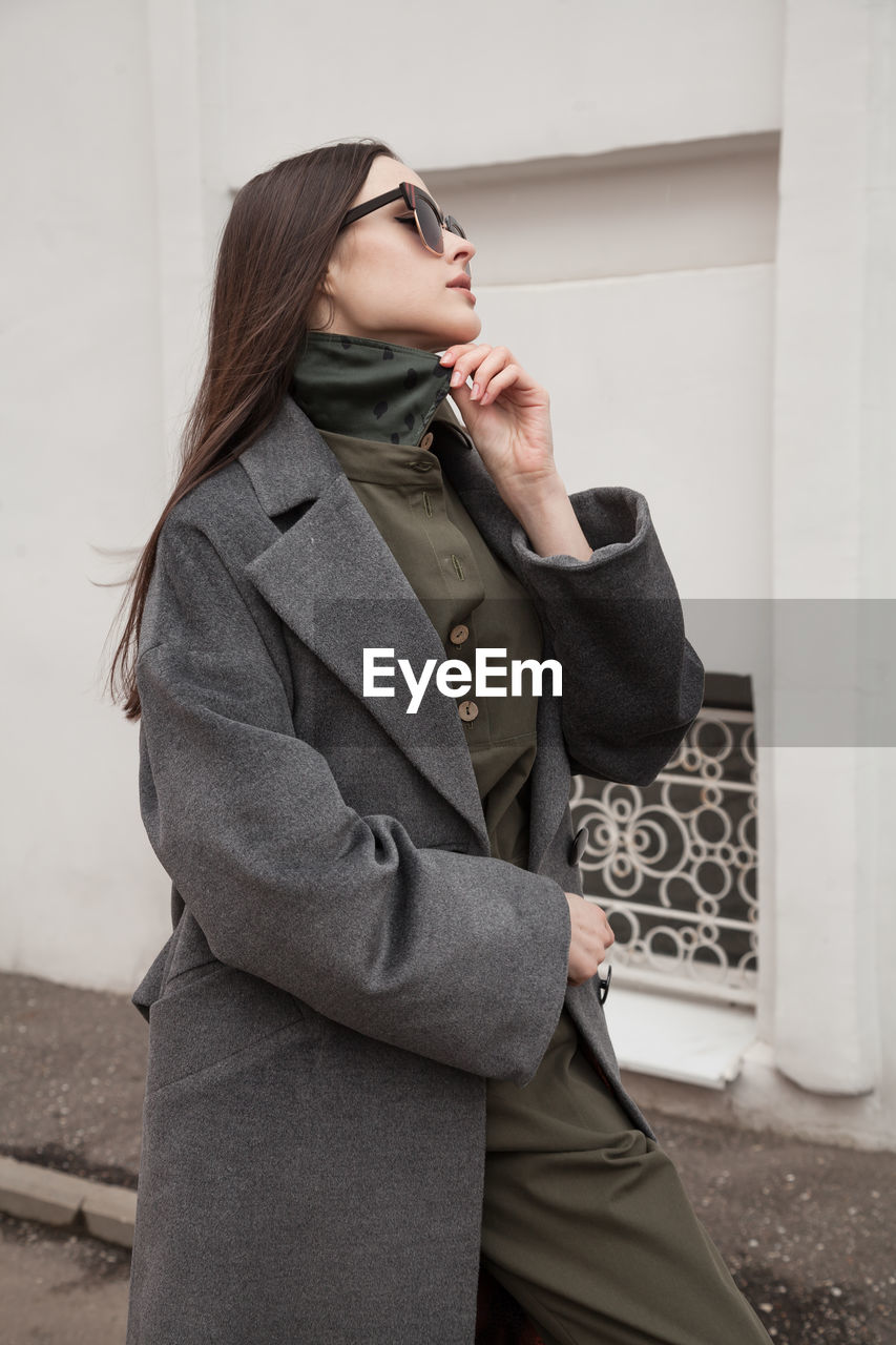 Close up side view postttait of young woman in grey coat standing against wall outdoors, outwear 