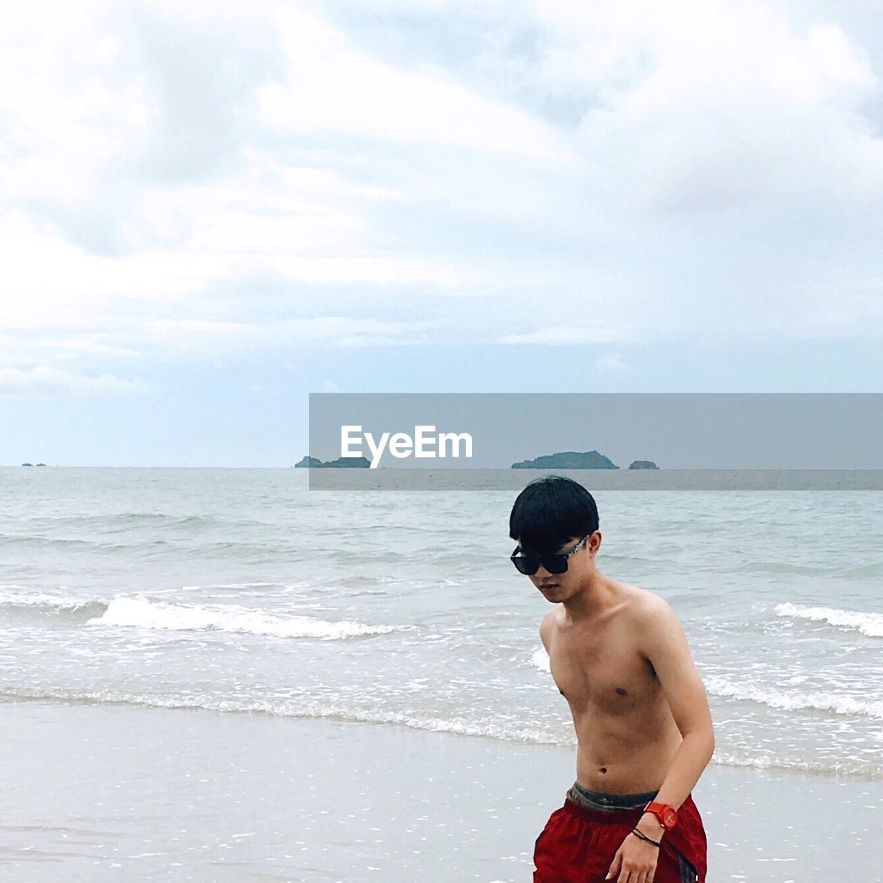 Shirtless young man walking on shore at beach against cloudy sky