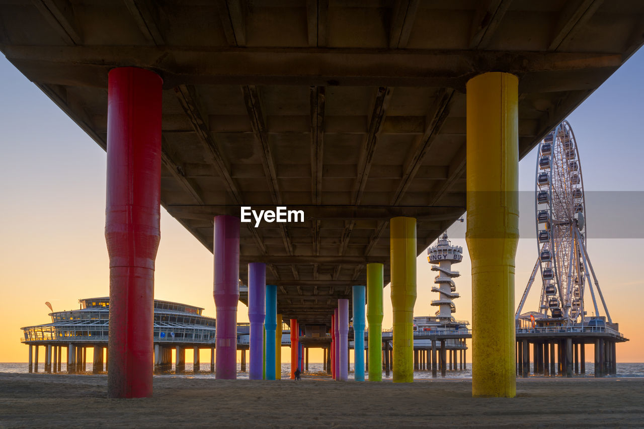 Multi colored poles under bridge against sky during sunset