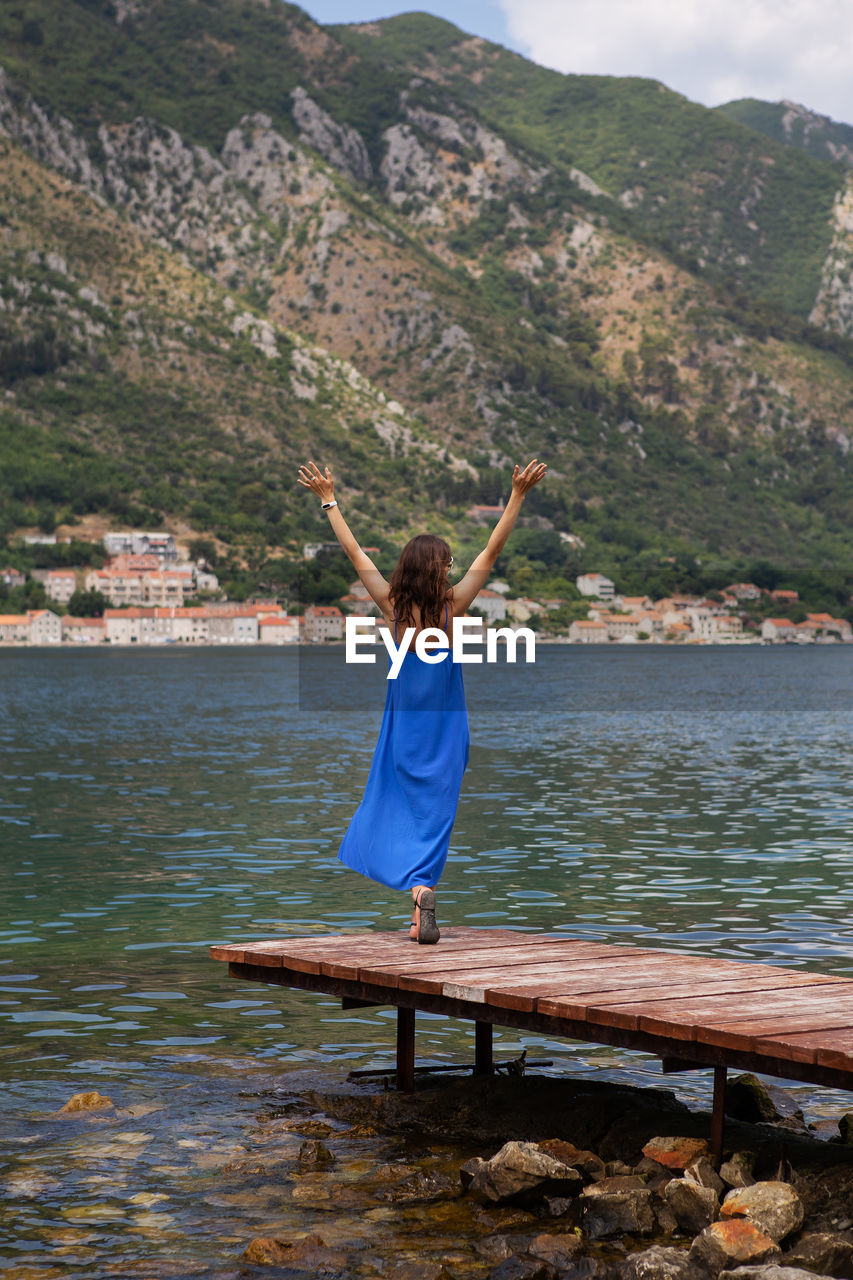 A girl in a dress stands with her hands up on the pier of the bay of kotor in montenegro.