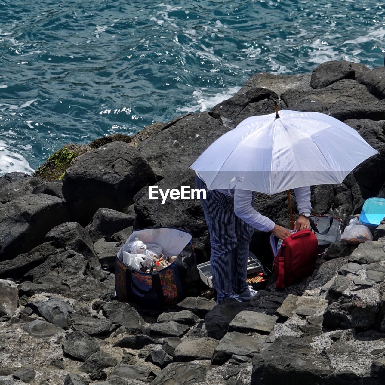 High angle view of man with bags and umbrella standing on rocks by sea