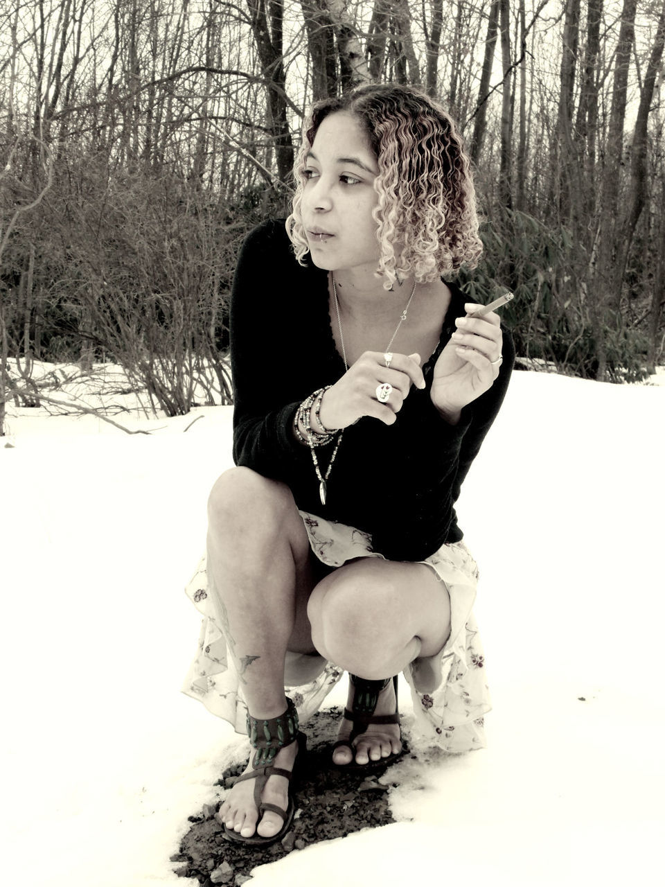 Young woman smoking while crouching on snowy field against bare trees