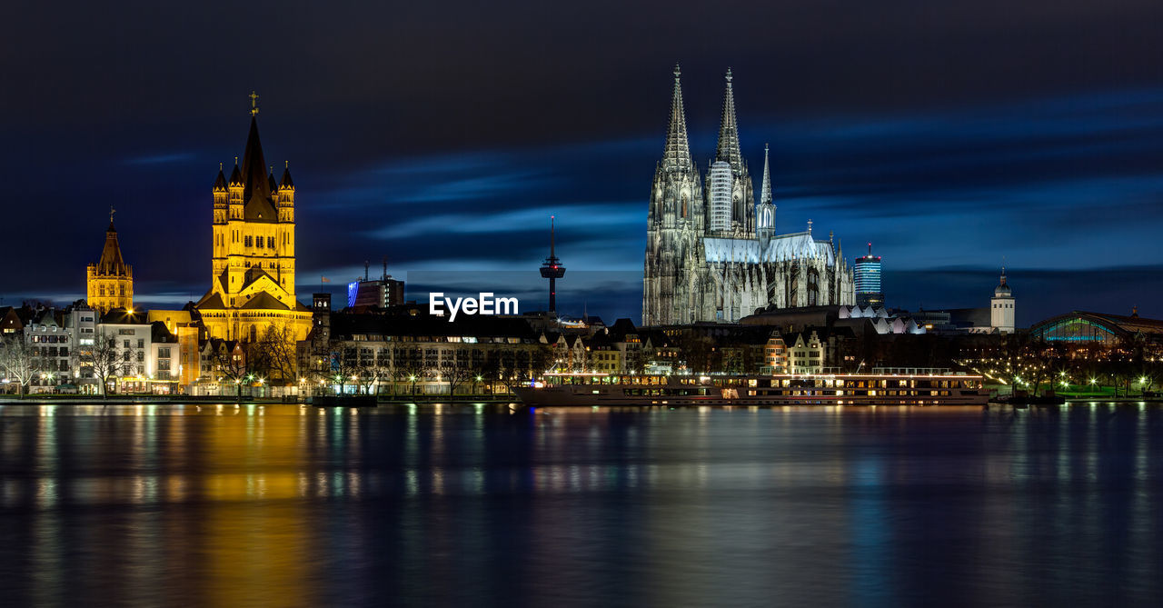 Illuminated buildings and cologne cathedral by rhine river in city at night