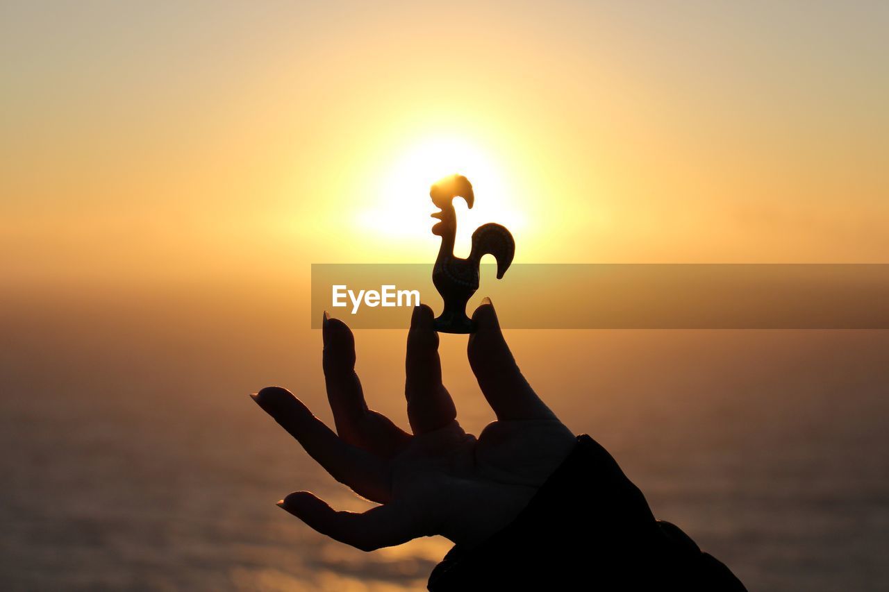 Cropped image of person holding silhouette rooster figurine by sea against sky during sunset
