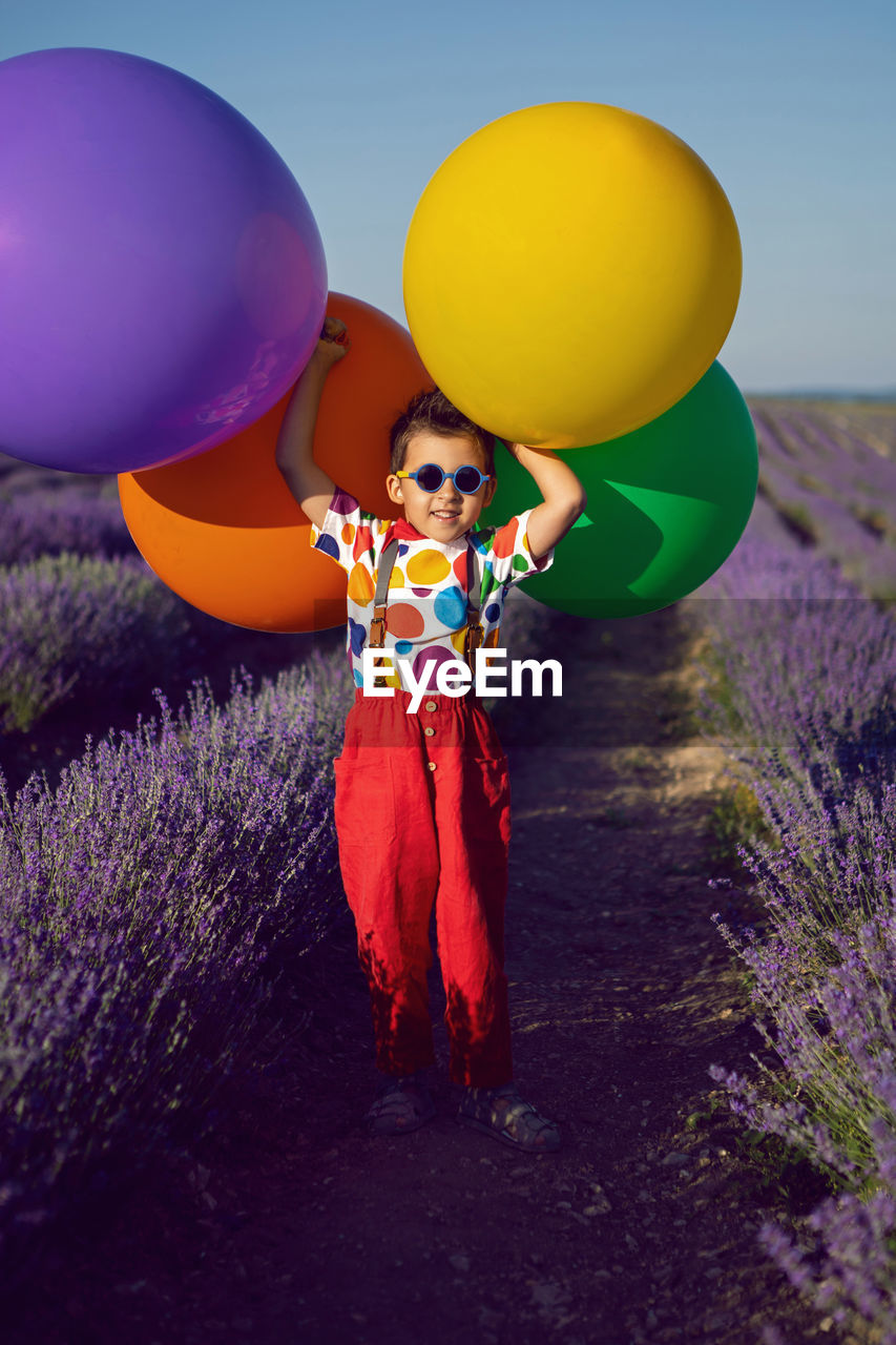 balloon, multi colored, one person, nature, flower, plant, celebration, sky, child, helium balloon, childhood, purple, women, adult, toy, portrait, full length, fun, female, land, happiness, emotion, yellow, smiling, helium, standing, bunch, blue, landscape, event, outdoors, holding, flowering plant, looking at camera, clothing, field, environment, cute, tradition, cartoon, front view