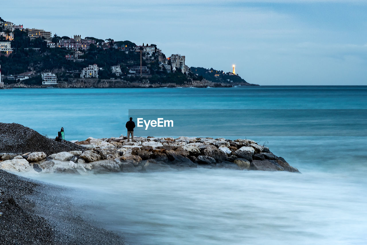 Rear view of man standing on rocks by sea against sky