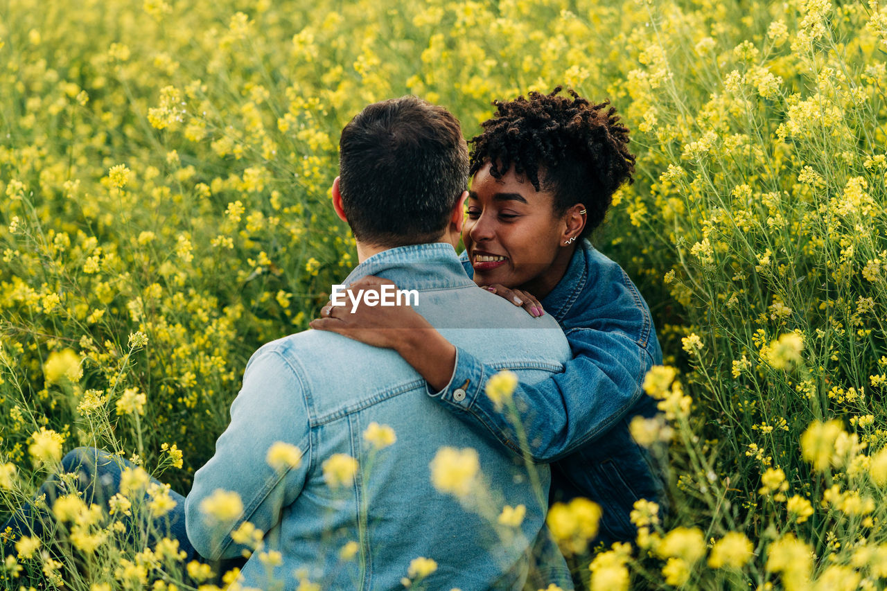 Loving young multiracial couple in casual clothes looking at each other while sitting in lush blooming meadow during romantic date on sunny day