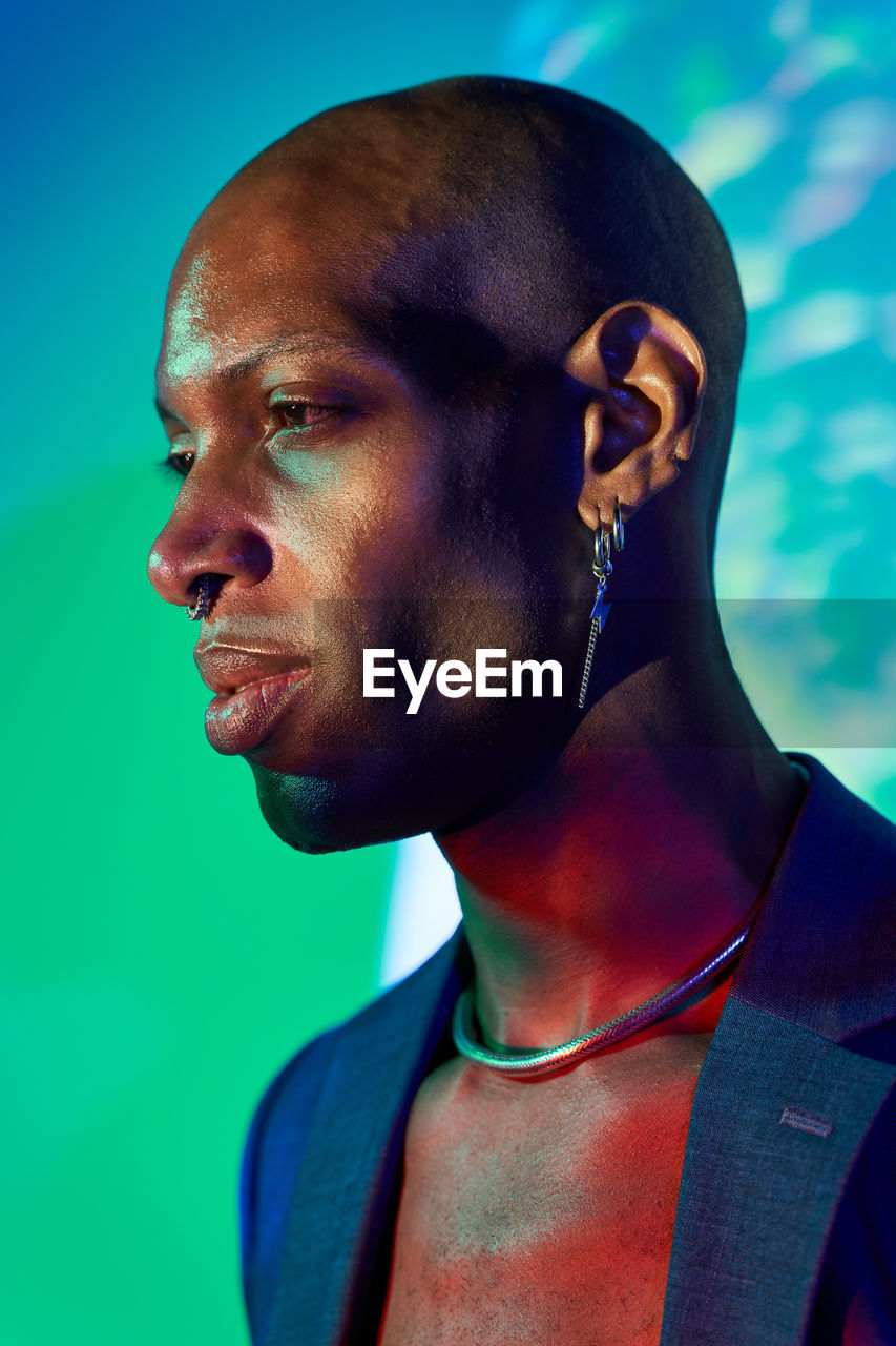 Confident bald african american male model with necklace and earring in trendy outfit standing at wall with glowing lights in modern studio looking away