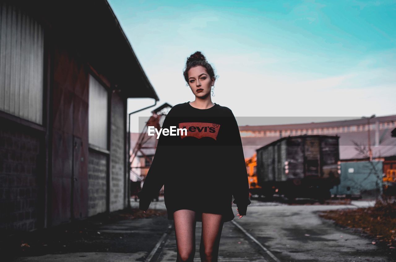 PORTRAIT OF YOUNG WOMAN STANDING BY BUILDING AGAINST SKY