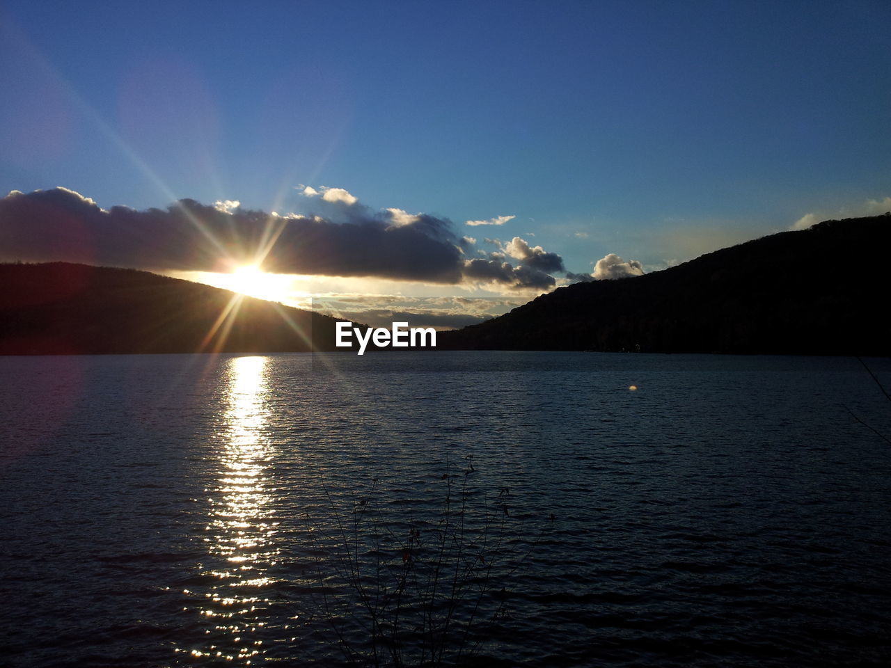 Sun shining through silhouette mountains by lake