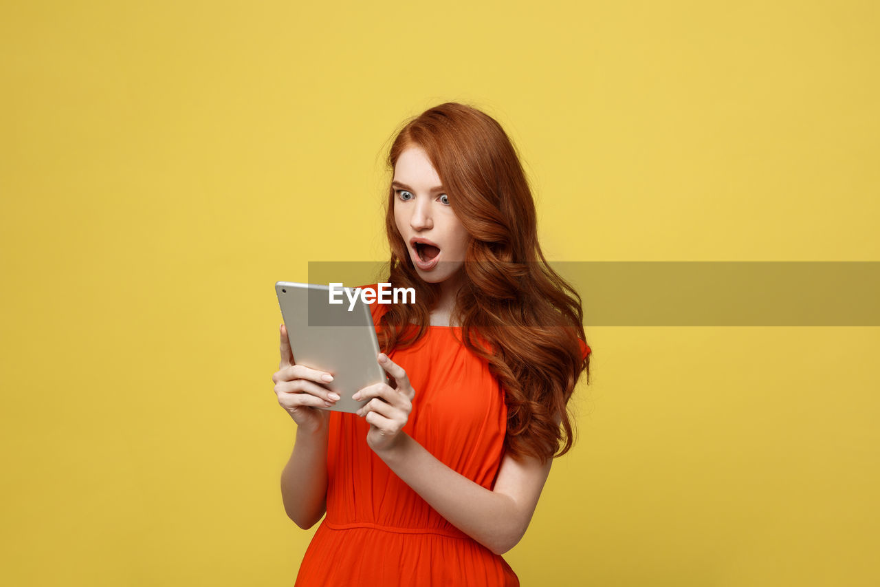 Young woman with digital tablet standing against yellow background