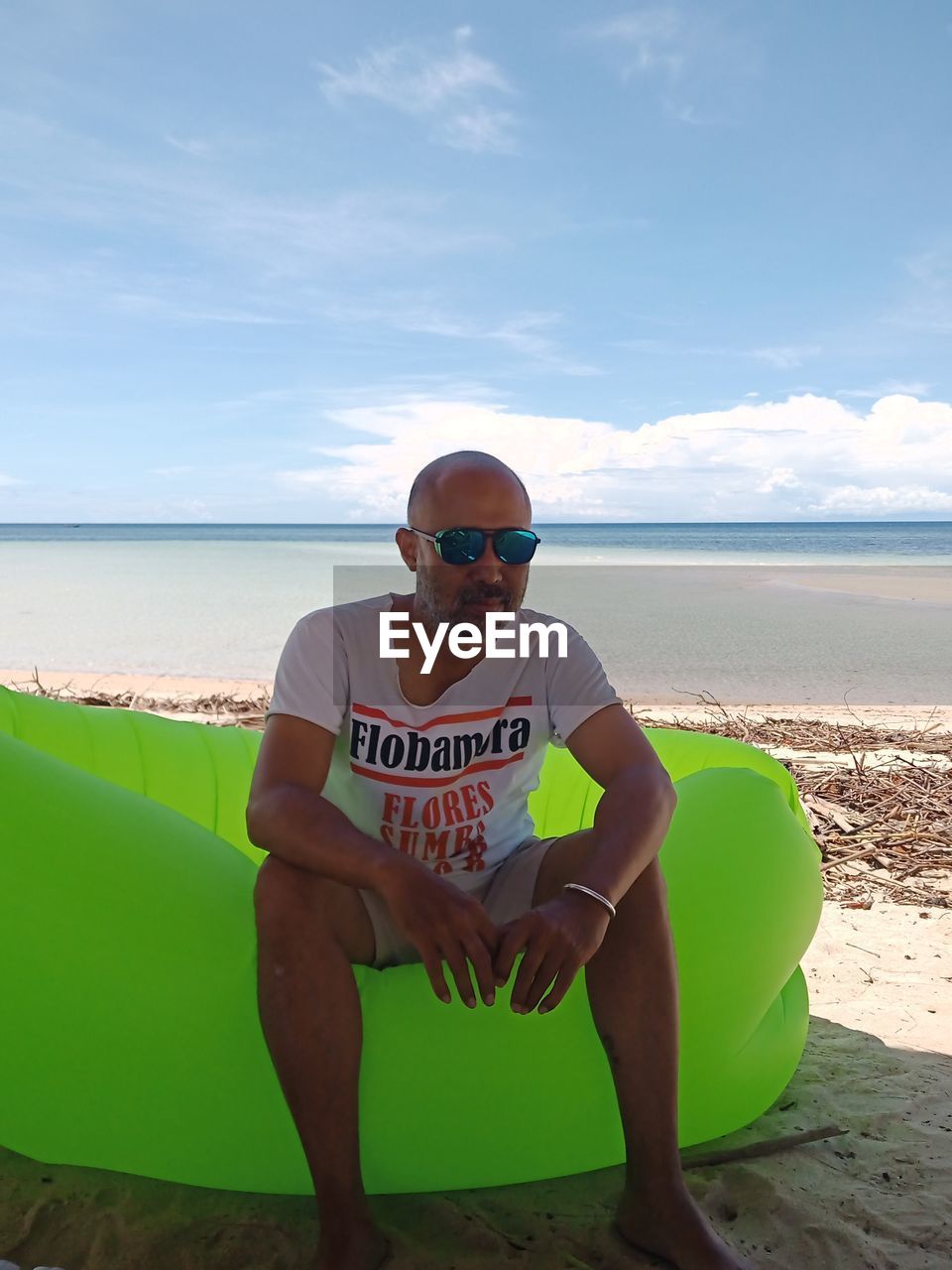 beach, water, sea, land, one person, vacation, sky, nature, full length, sunglasses, leisure activity, adult, sitting, glasses, front view, sand, fashion, trip, holiday, day, men, lifestyles, horizon over water, portrait, surfing equipment, cloud, young adult, sports, casual clothing, horizon, relaxation, looking at camera, outdoors, sun tanning, summer, clothing, travel, text, surfboard