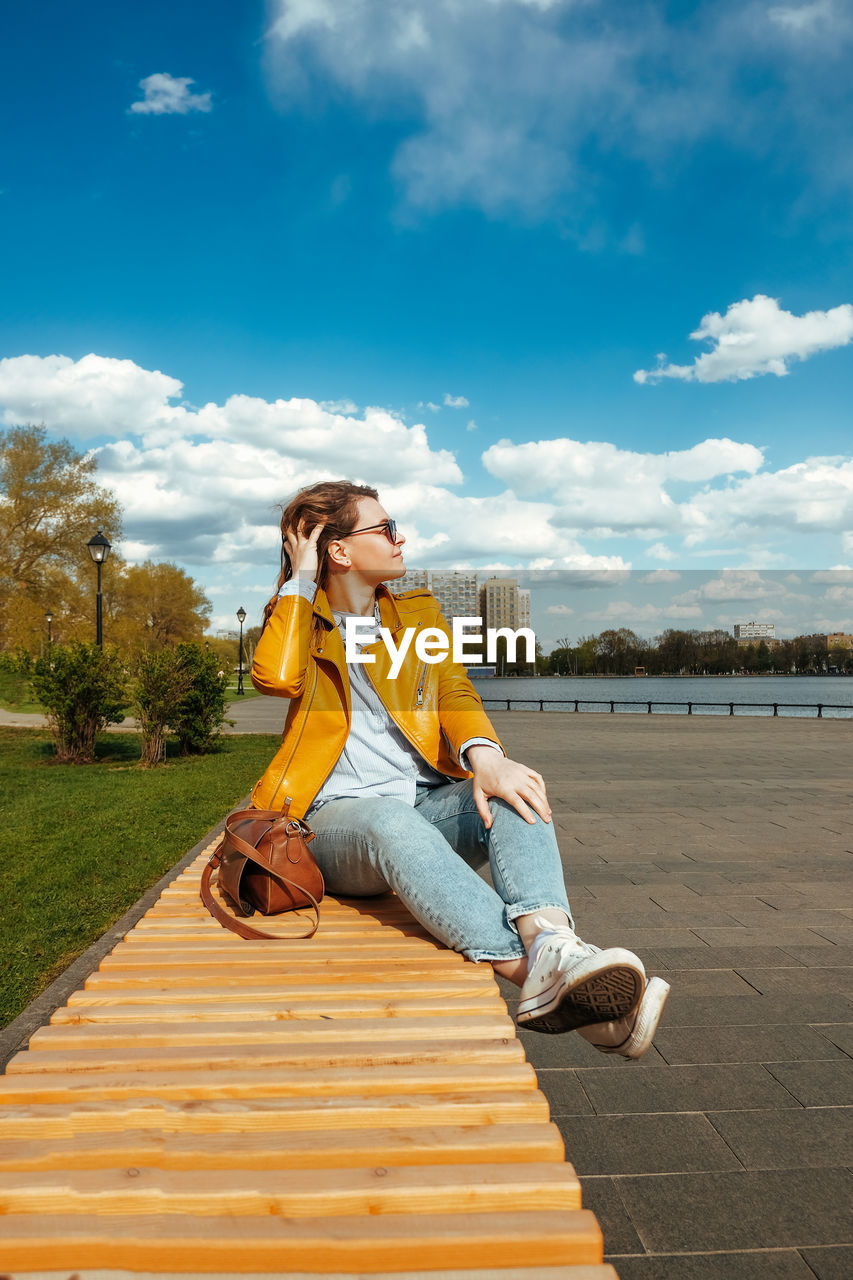 Young woman stylishly dressed in white sneakers, jeans sitting on street near pond. travel lifestyle