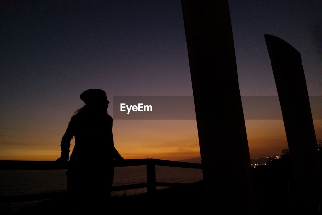 Silhouette of woman standing by railing against sky at sunset