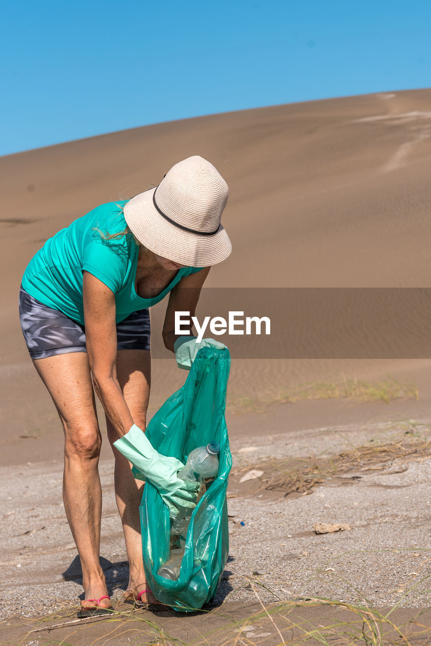 Unrecognizable adult woman in a white hat and green gloves picking up a plastic bottle on the sand.