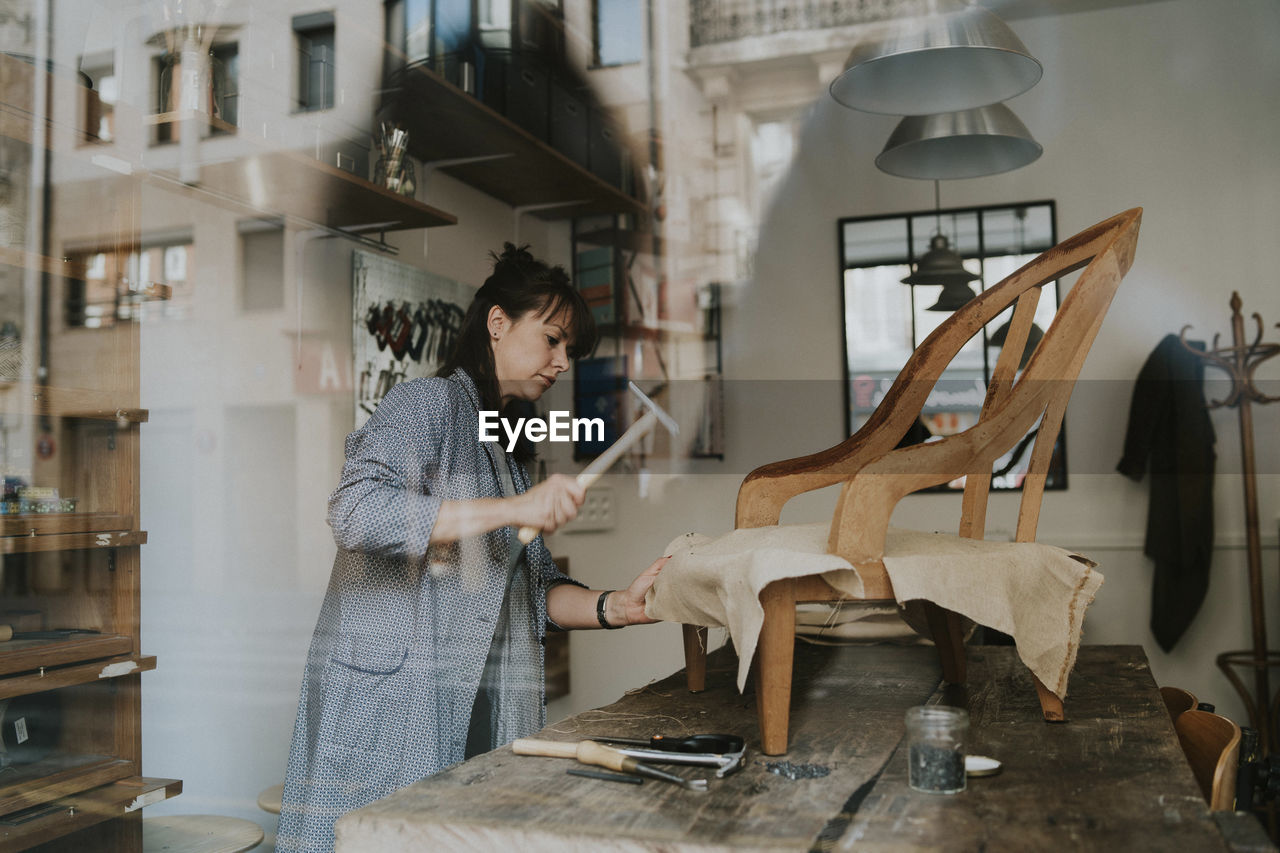 Female upholsterer making wooden chair on table in workshop seen through window