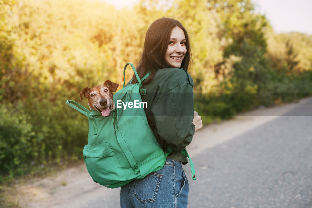 A smiling girl is holding a green backpack on her shoulder, from which a cute dog looks out. 