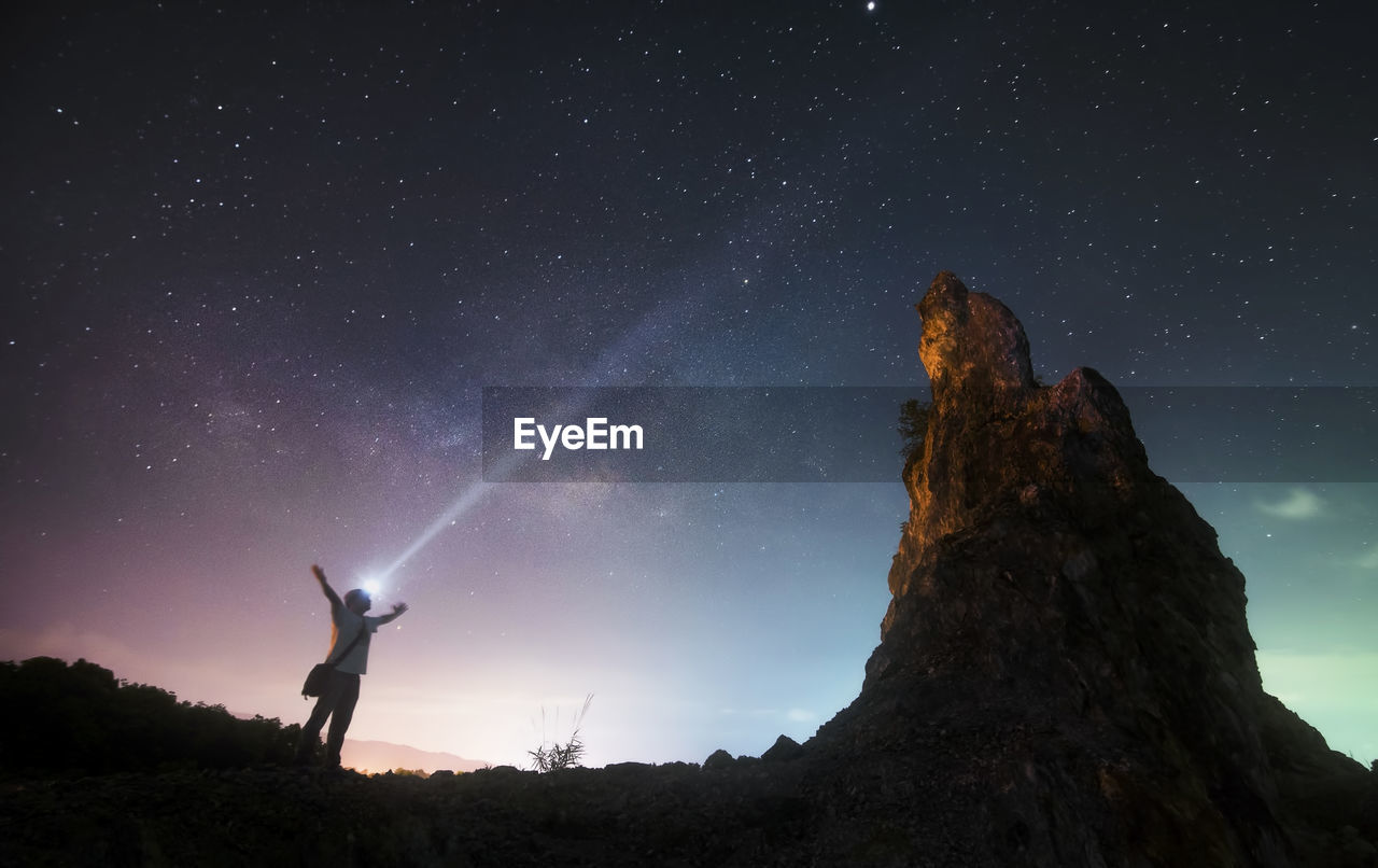 Low angle view of man with flashlight standing by rock formation against sky