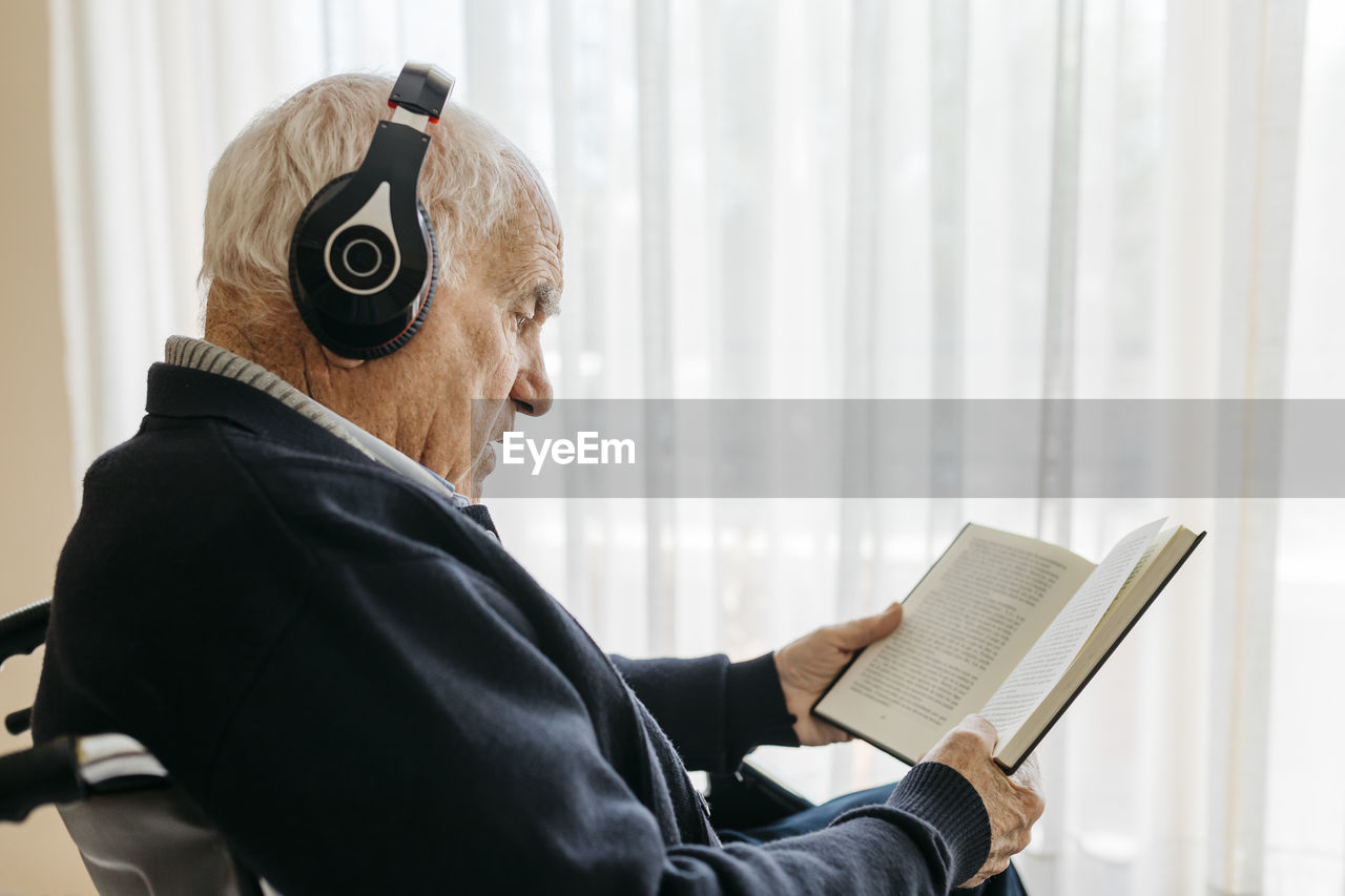 Senior man sitting in wheelchair reading a book while listening music with headphones