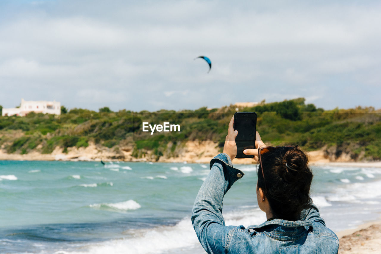 Rear view of woman photographing through mobile phone at beach