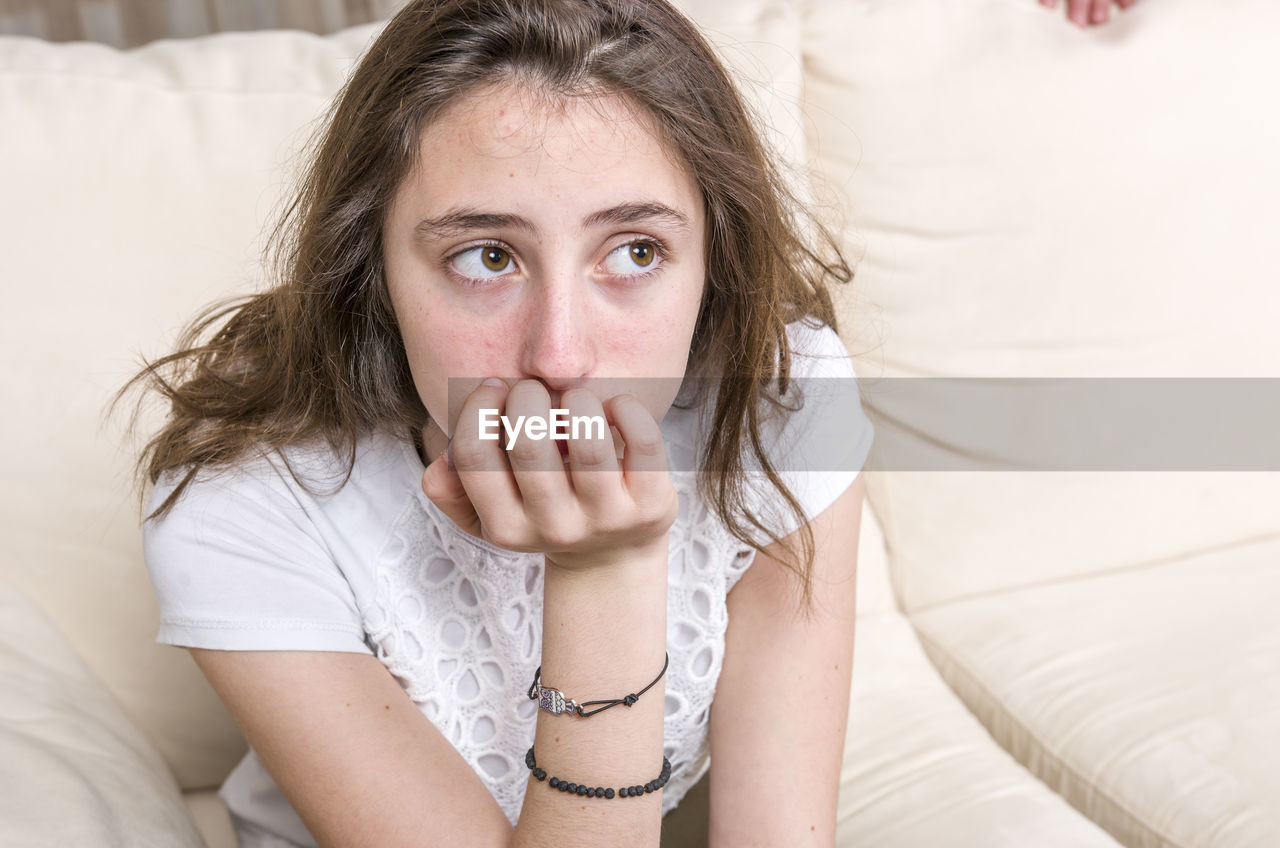 Thoughtful teenage girl with hand on chin sitting on sofa at home