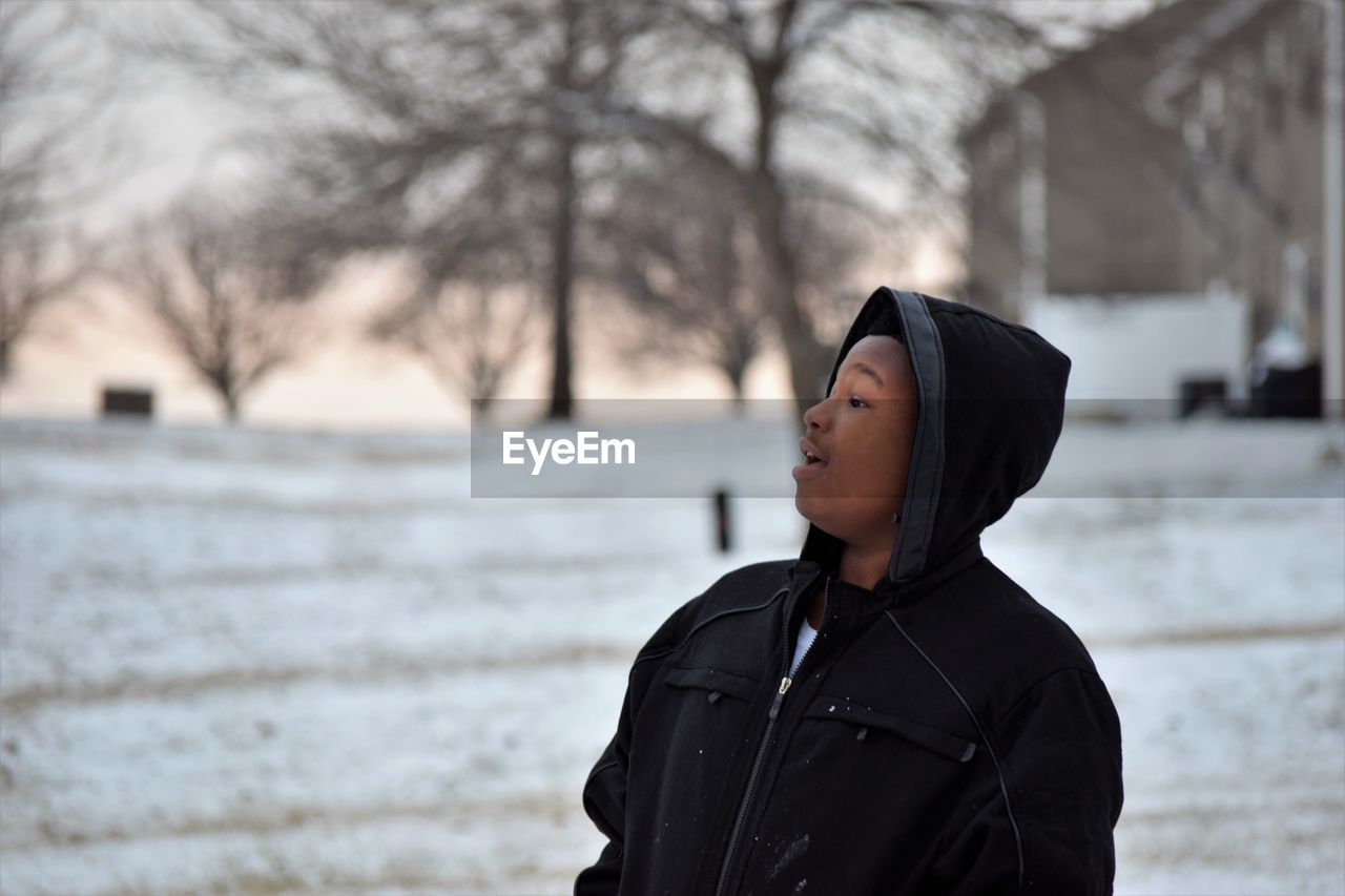 Man in hooded jacket and looking away while standing on snow covered field