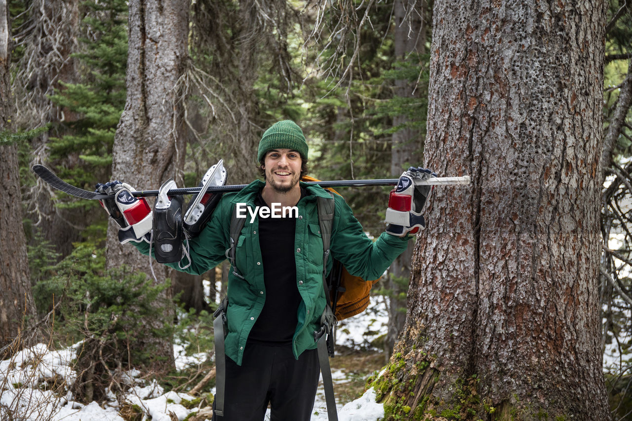 Cheerful young man with hockey stick and ice skates on shoulders smiling and looking at camera while standing near trees in snowy forest