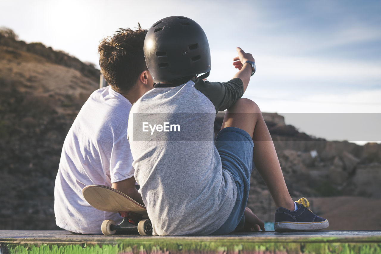 Rear view of boy sitting with friend on retaining wall