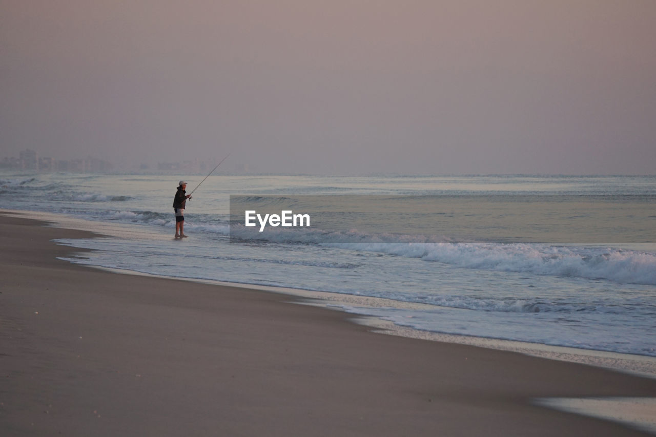 Man fly-fishing while standing on shore at beach during sunset