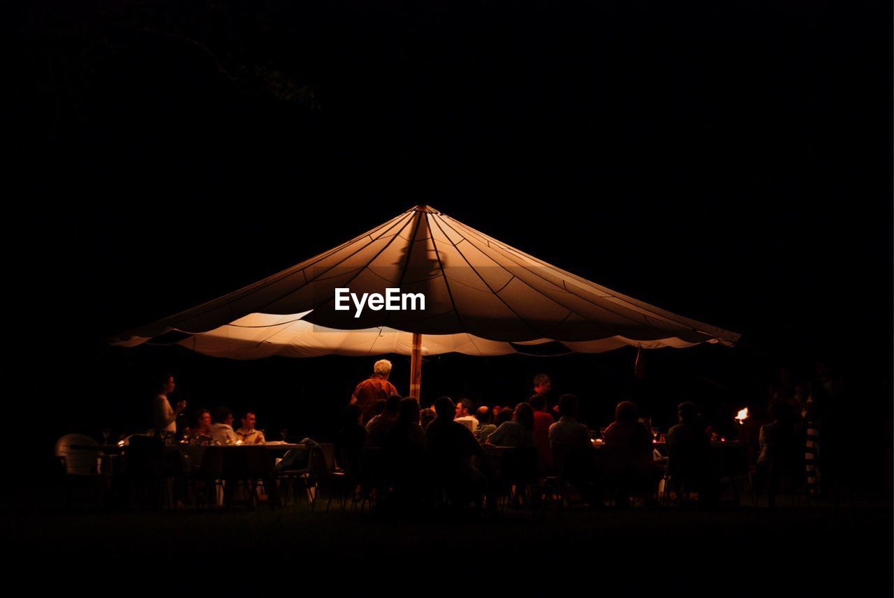 People enjoying dinner at illuminated parasol against sky at night