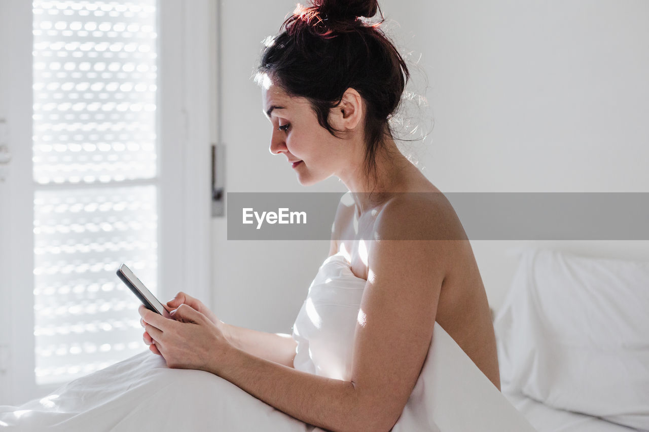 Side view of young woman using mobile phone on bed