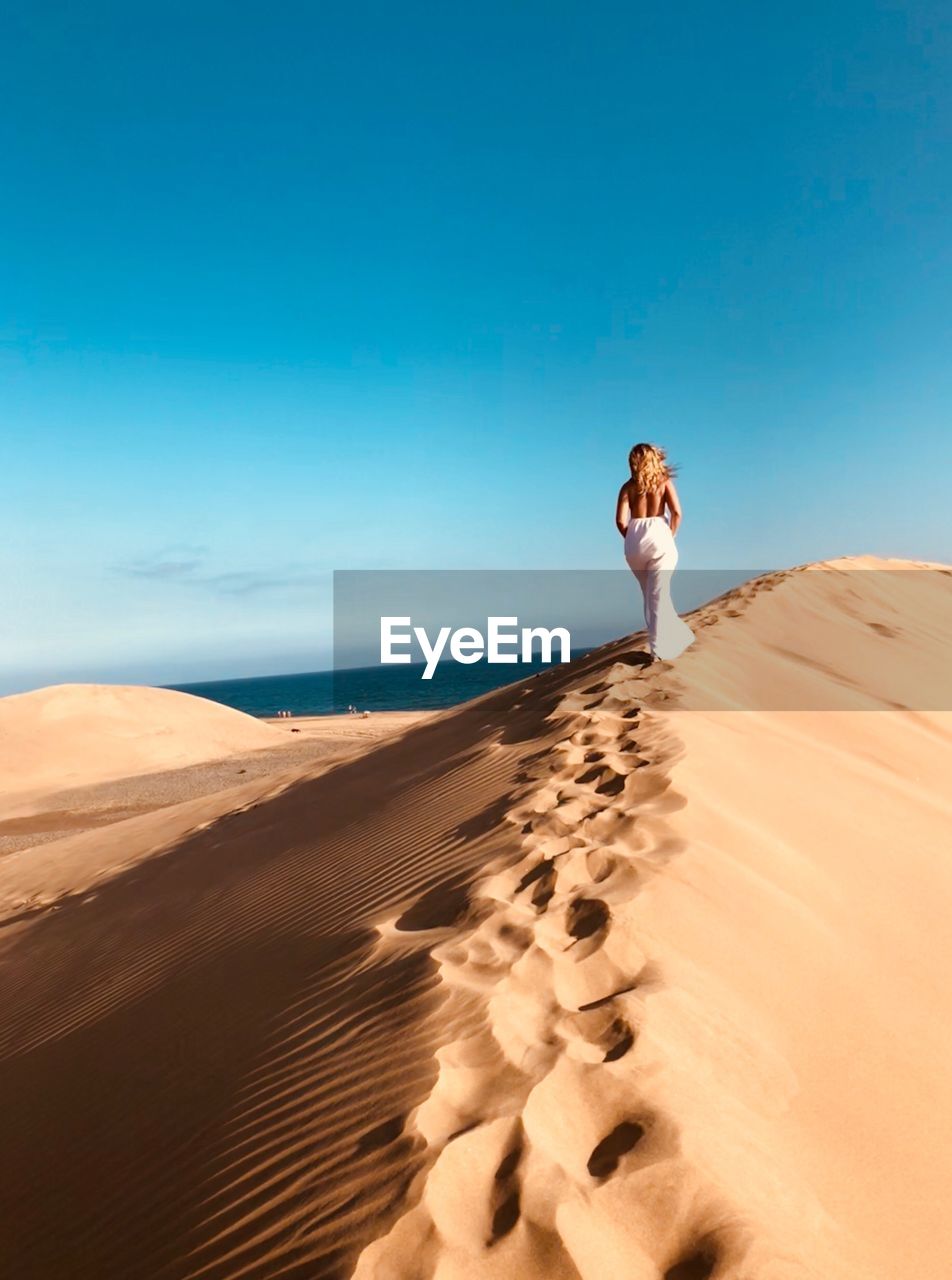 Rear view of woman walking on sand dune in desert against blue sky