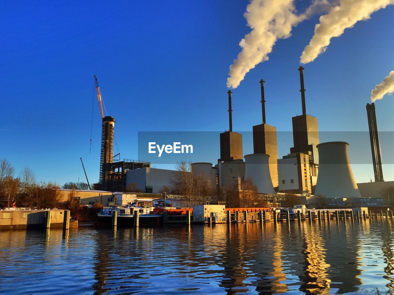 Power station on riverbank emitting smoke against clear blue sky