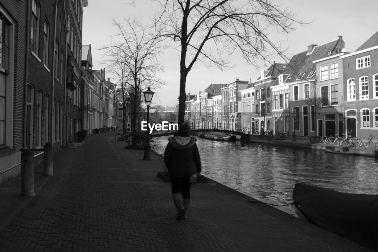 REAR VIEW OF MAN WALKING ON CANAL AMIDST BUILDINGS