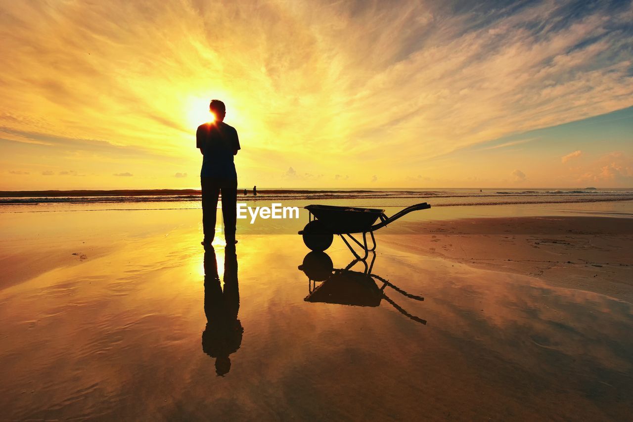 Silhouette man standing by wheelbarrow on shore at beach during sunset