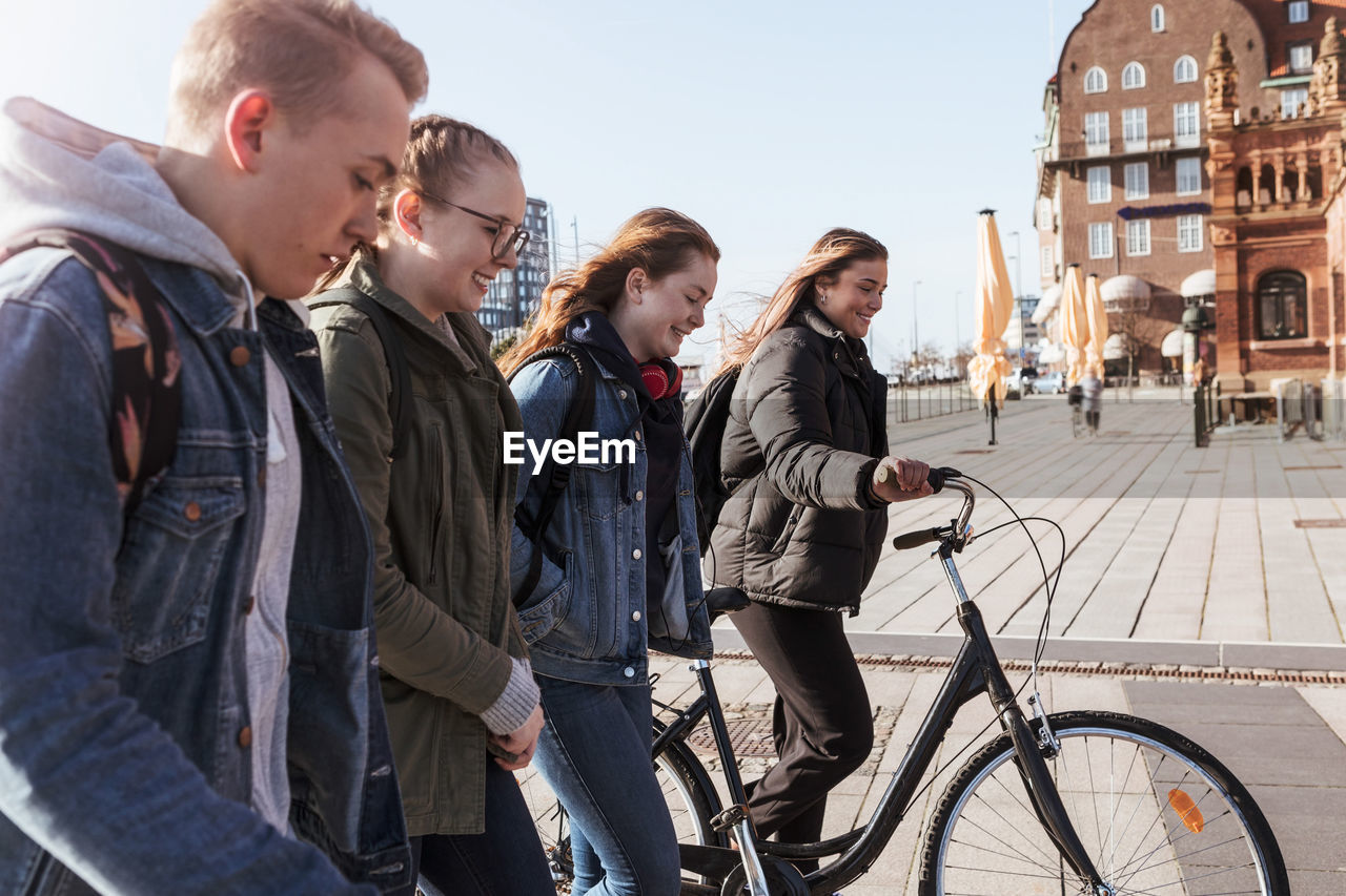 Side view of friends walking with teenage girl holding bicycle in city