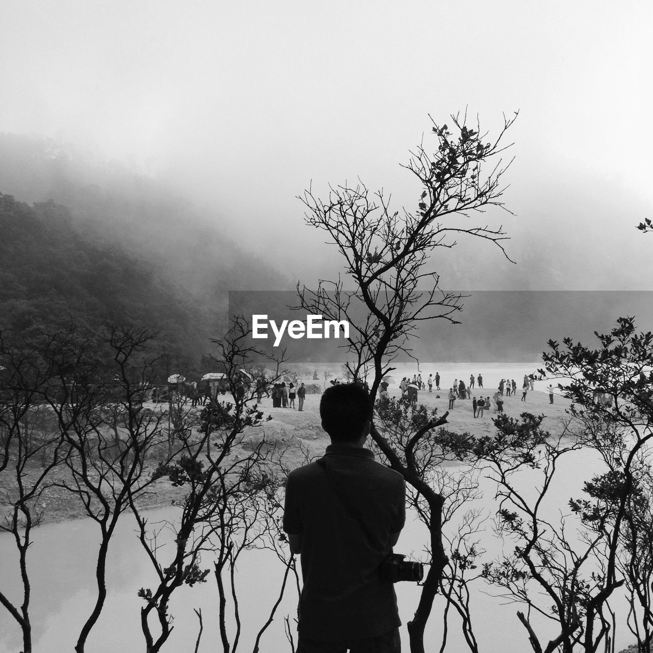 Rear view of photographer standing by river and tree mountain against sky during foggy weather