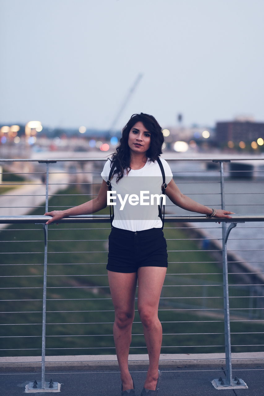 Portrait of mid adult woman standing by railing on footbridge against sky during sunset