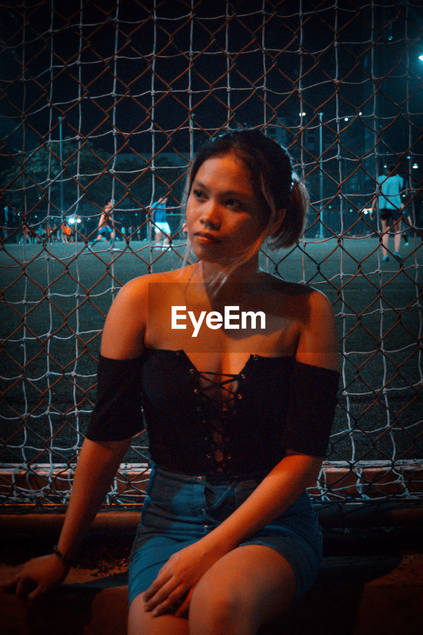 YOUNG WOMAN LOOKING AWAY WHILE SITTING AT CHAINLINK FENCE