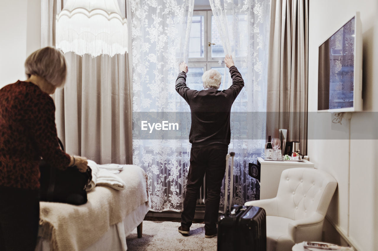 Senior man opening curtains while woman holding bag in hotel room