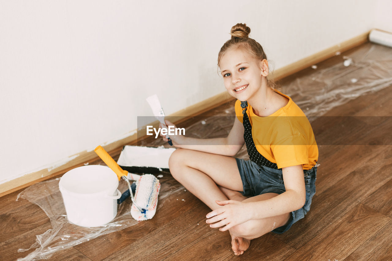 A girl in a denim overalls and a yellow t-shirt helps to paint the walls in an apartment white.