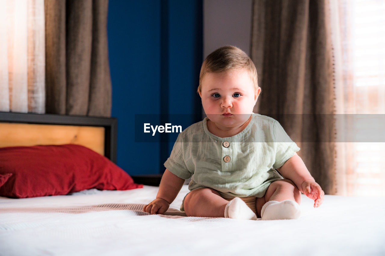 PORTRAIT OF CUTE BABY SITTING ON BED AT HOME