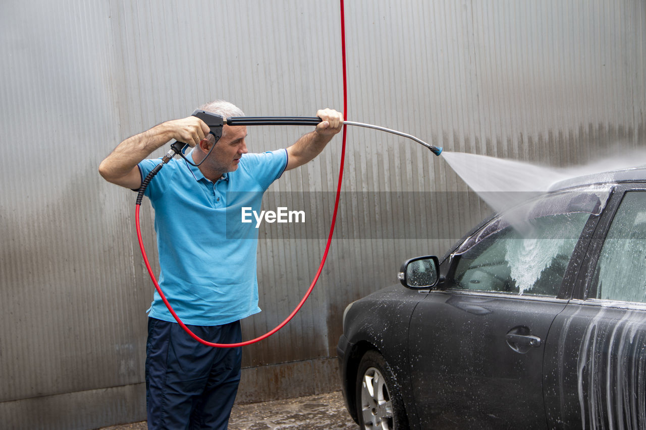 Mature man cleaning automobile with foam shampoo chemical detergents during carwash self service