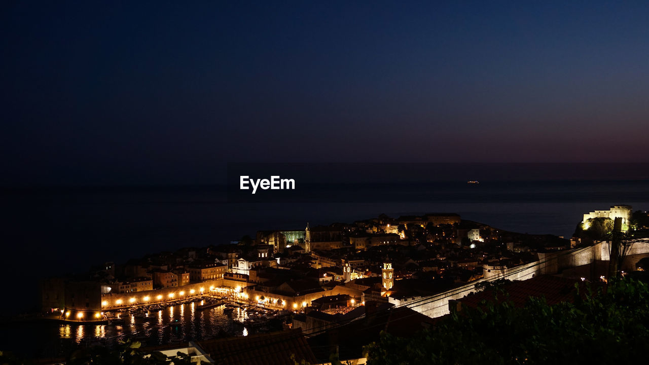 HIGH ANGLE VIEW OF ILLUMINATED CITY BY SEA AGAINST CLEAR SKY AT NIGHT