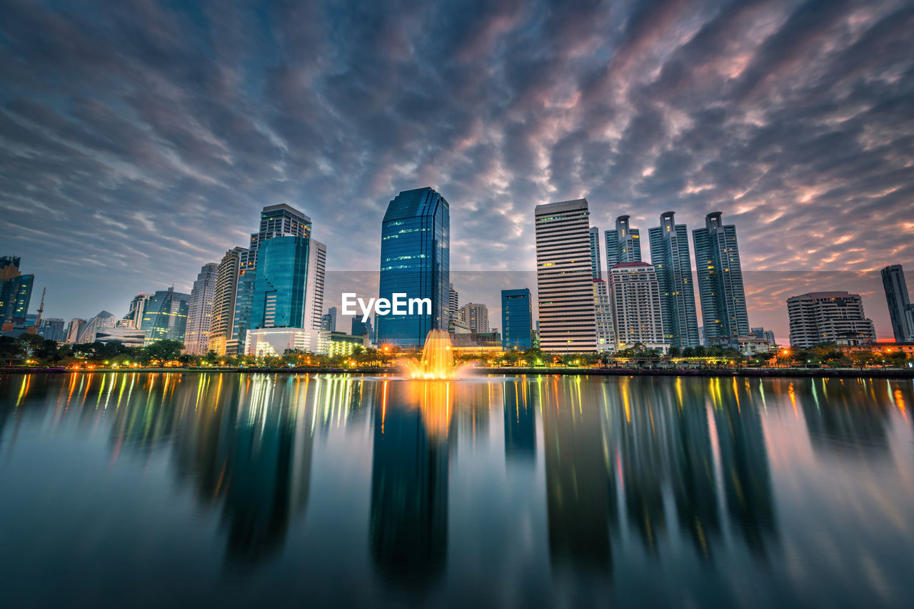 Reflection of illuminated buildings in city against sky during sunset