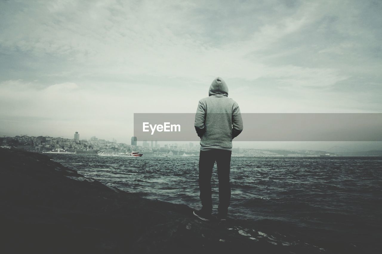 Rear view of man standing at shore against cloudy sky