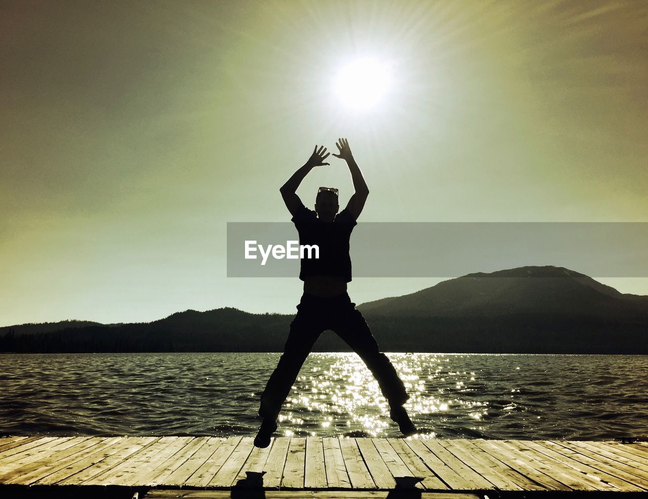 Silhouette of man jumping with arms raised on pier over lake at sunset