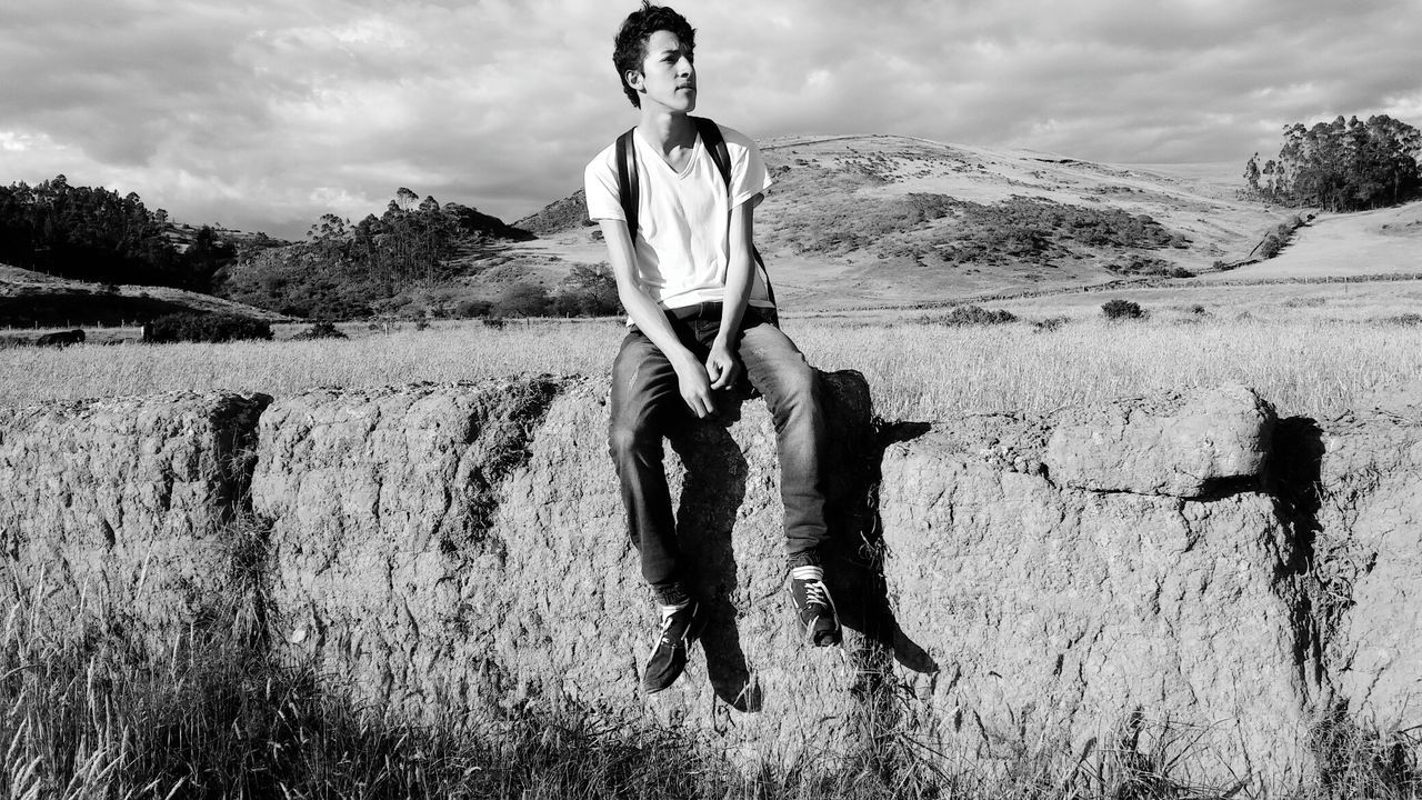 Young man sitting on rock by grassy field against hill